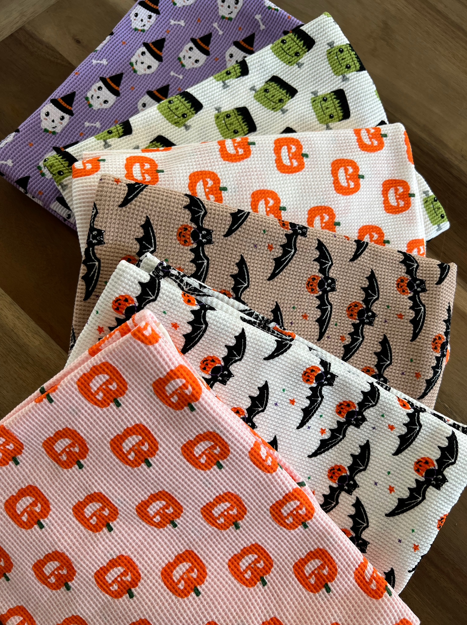 Colorful Halloween-themed kitchen towels featuring pumpkins, ghosts, and bats stacked on a wooden surface.
