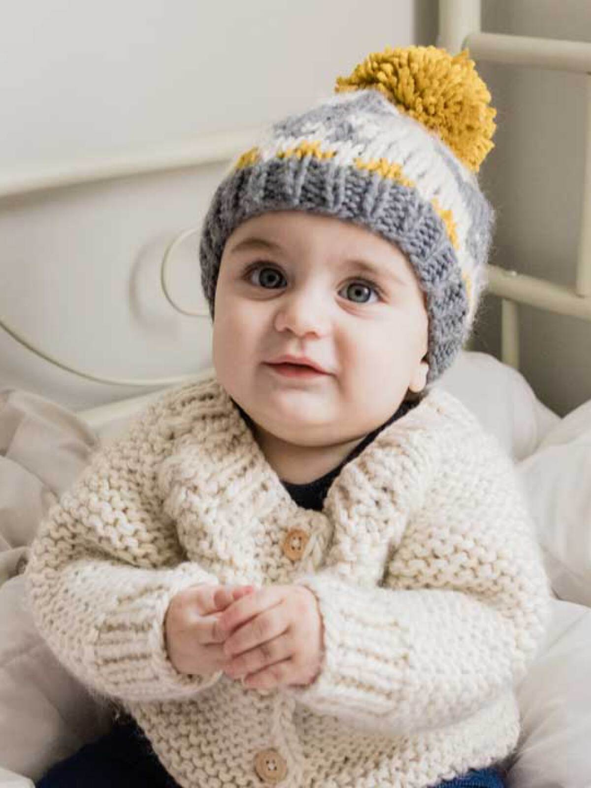 Smiling baby in a cozy sweater and knitted hat, sitting on a soft bed, with hands clasped together.