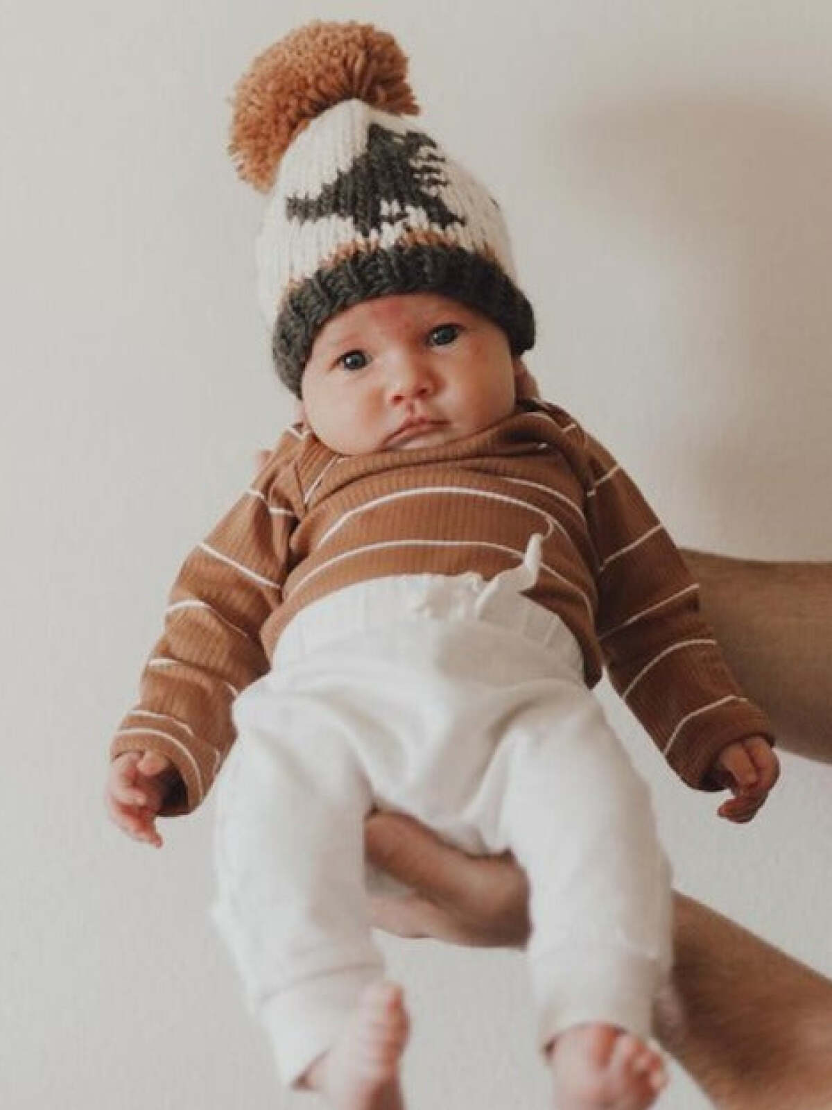 Baby in a cozy brown striped onesie and a patterned knit hat, held by an adult's hand against a neutral wall.