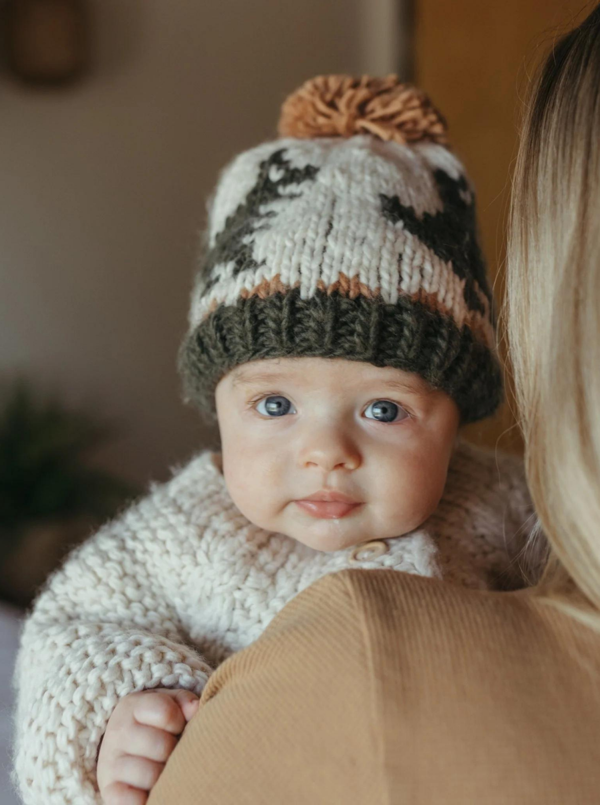 Baby wearing a knitted hat and cozy sweater, looking curiously at the camera while being held.