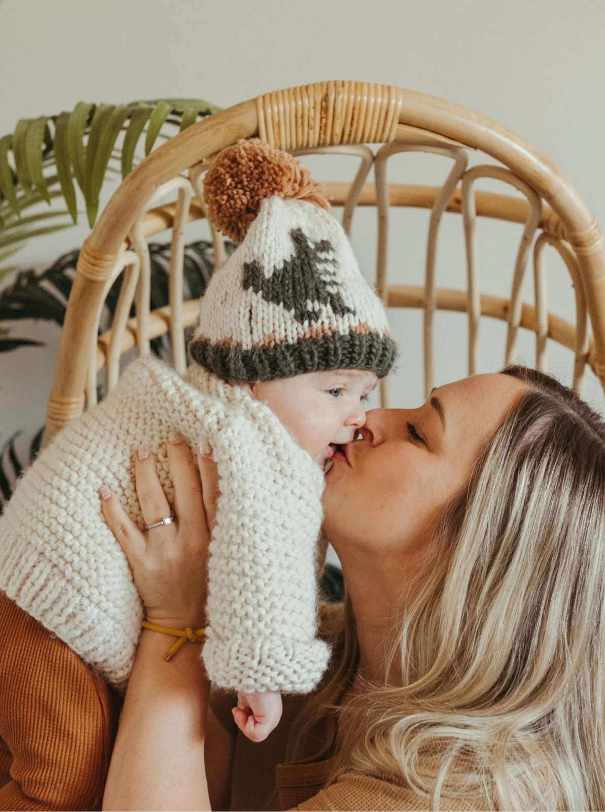 Mother kisses baby in cozy sweater and hat, seated in a rattan chair with greenery in the background.