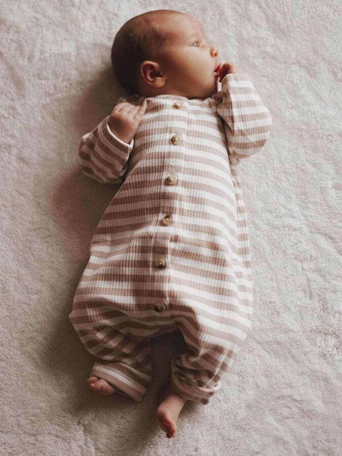 Baby in striped beige and white onesie, lying on a soft light-colored surface, looking thoughtfully to the side.
