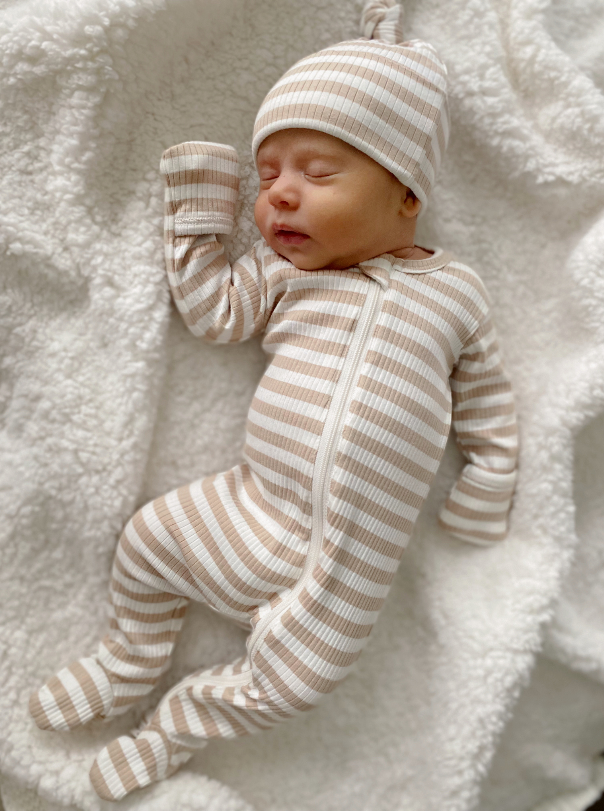 Sleeping newborn dressed in a striped pajama and hat, resting on a fluffy white blanket.