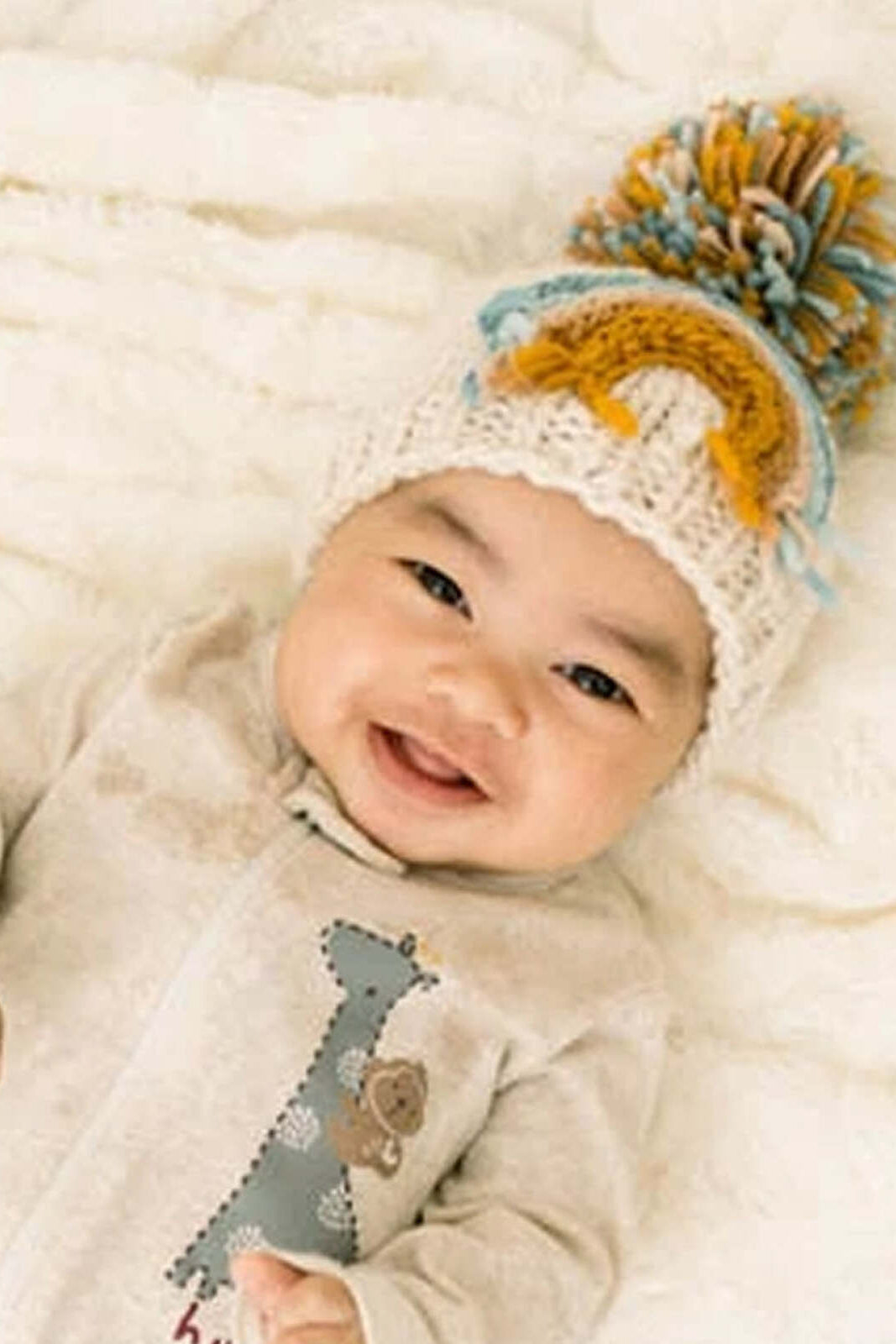 Happy baby wearing a knitted hat with pom-poms, laying on a soft blanket.