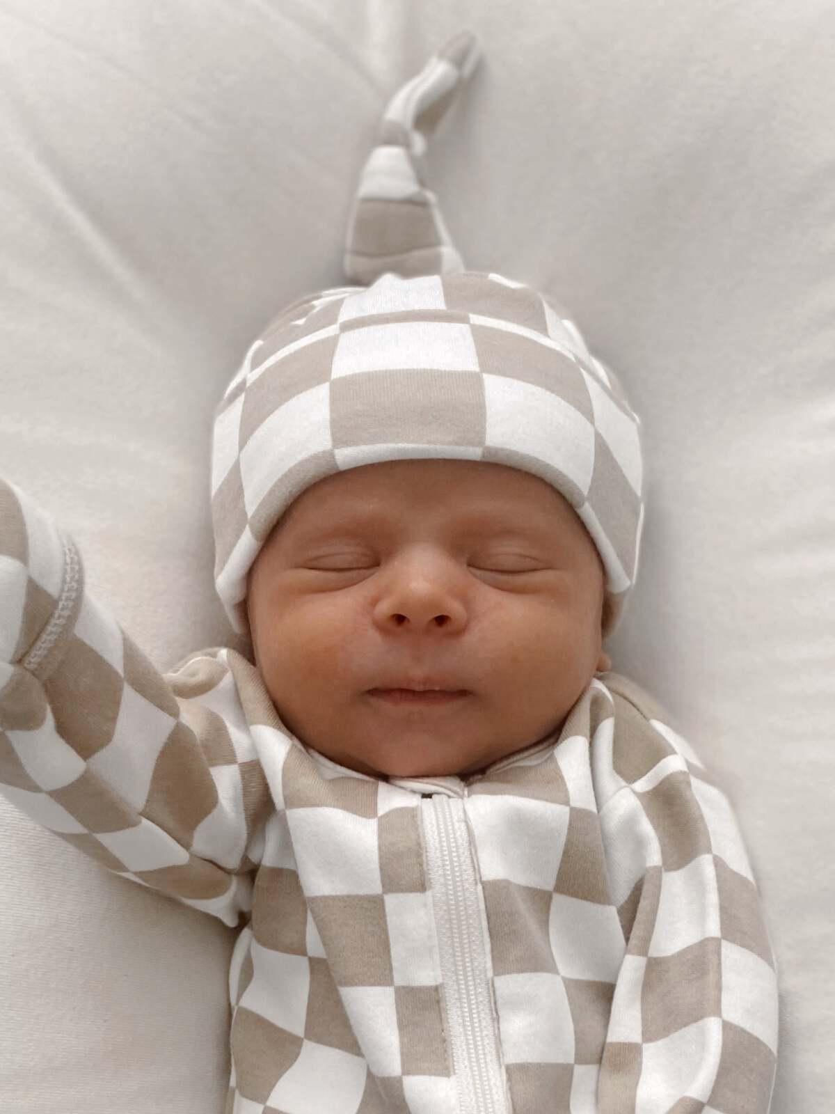 Newborn baby in a checkered outfit and hat, peacefully sleeping on a soft surface.