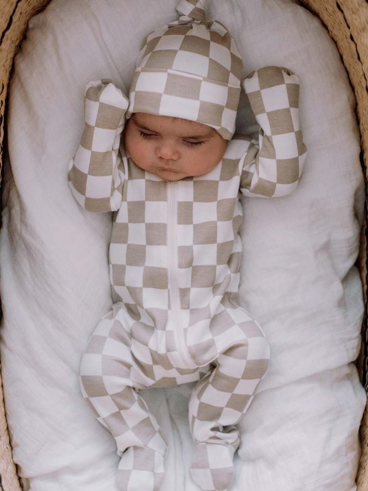 Baby in a checkered outfit and hat, lying on a white blanket in a cozy setting.