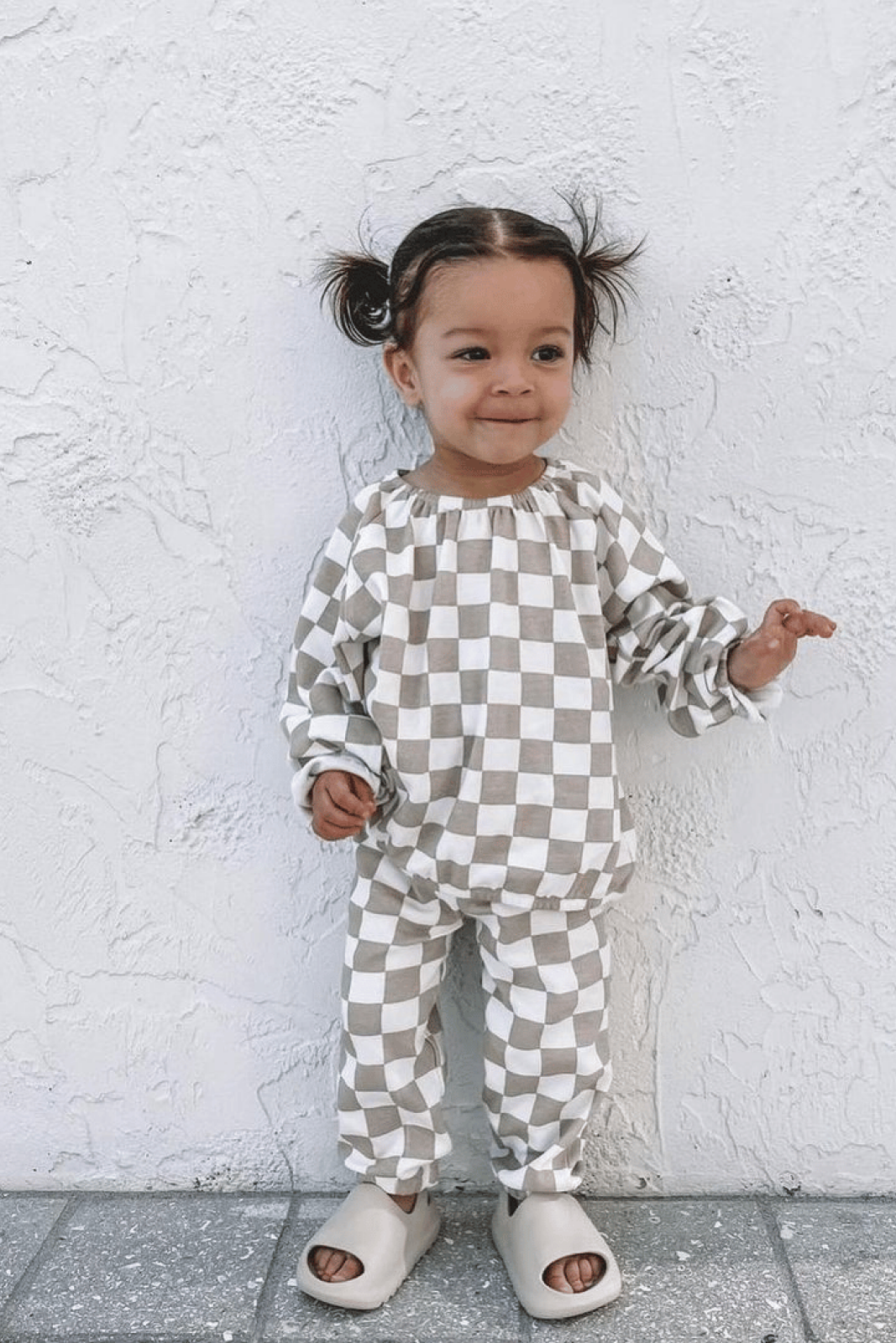 Smiling toddler in checkered outfit standing against a textured white wall, wearing light shoes.