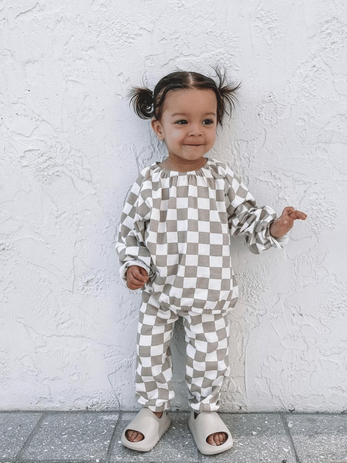 Smiling toddler in checkered outfit standing against a textured white wall, wearing light shoes.