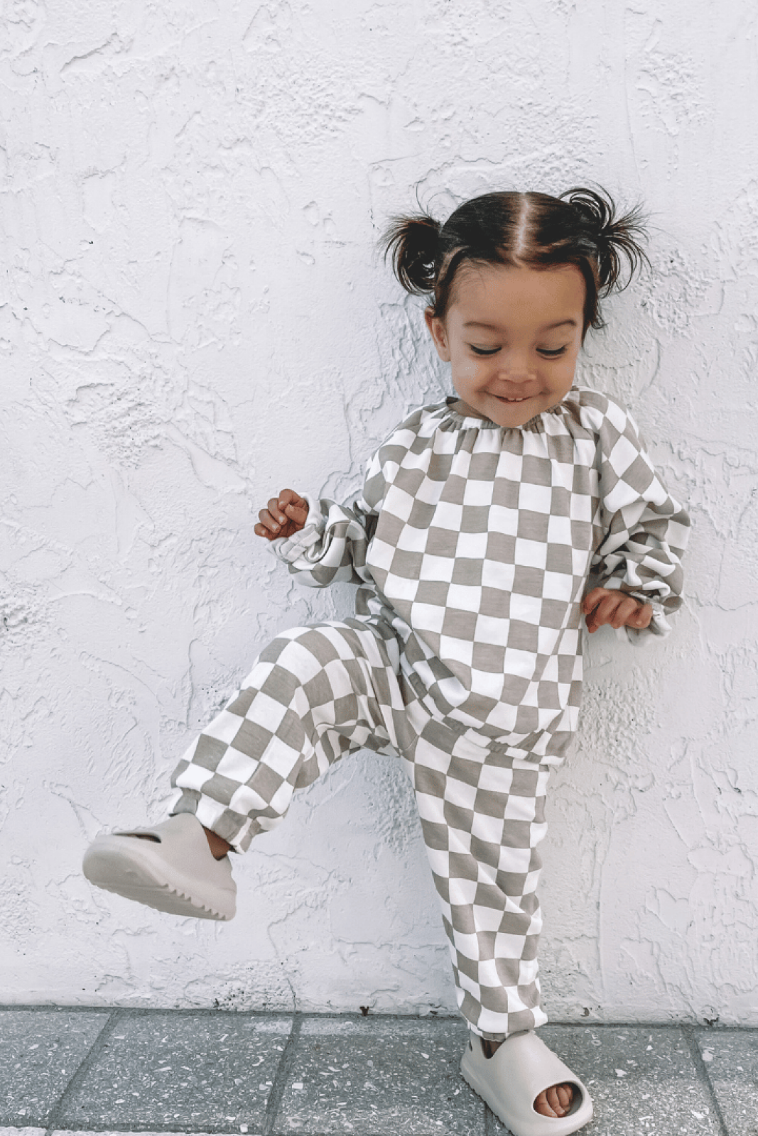 Toddler in checkered outfit playfully posing against a textured white wall.