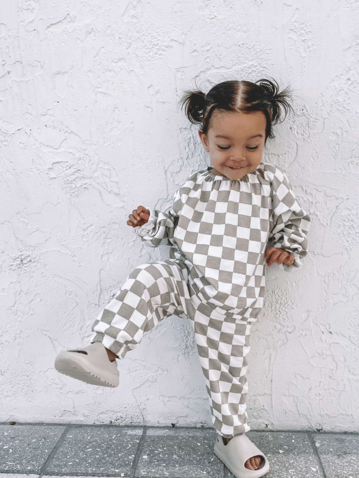 Toddler in checkered outfit playfully posing against a textured white wall.