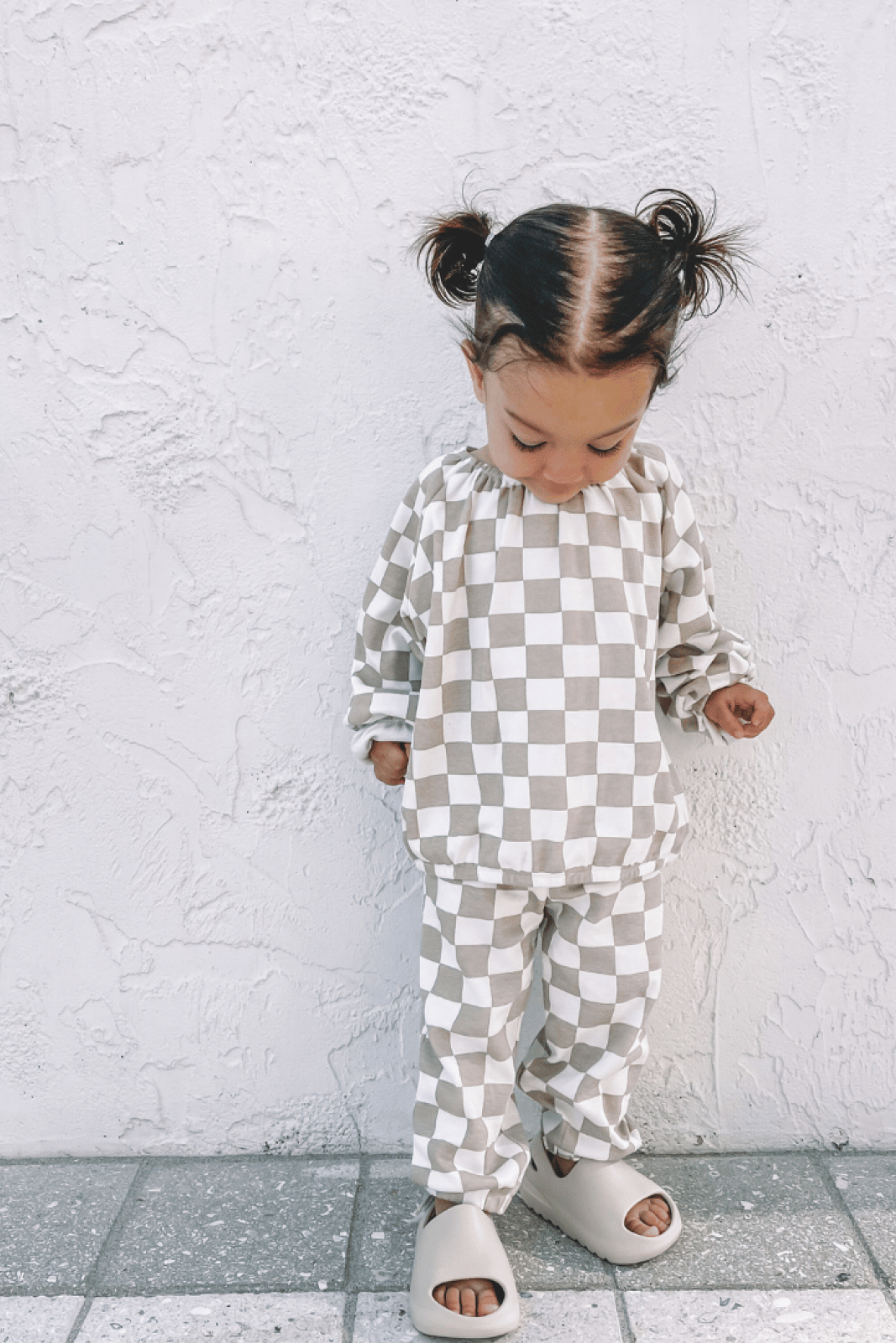Child in checkered outfit with pigtails, standing on a tiled floor against a textured white wall.