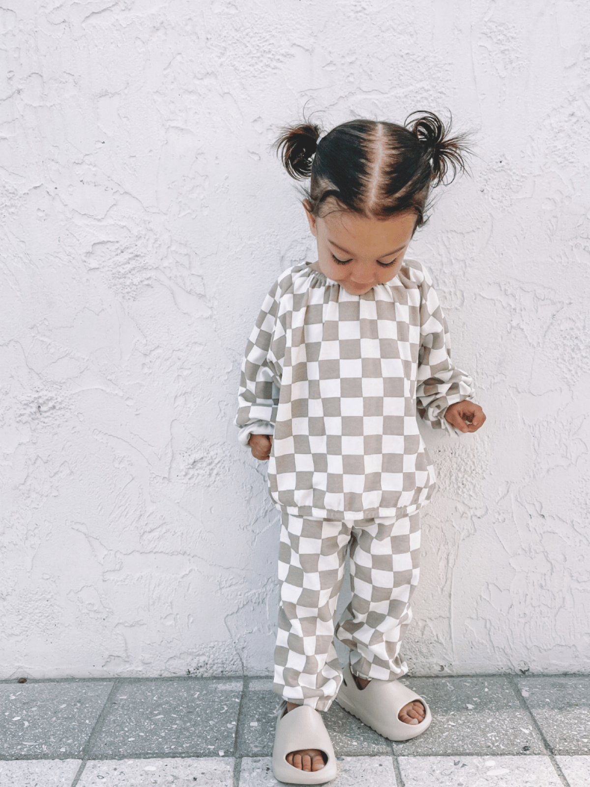 Child in checkered outfit with pigtails, standing on a tiled floor against a textured white wall.