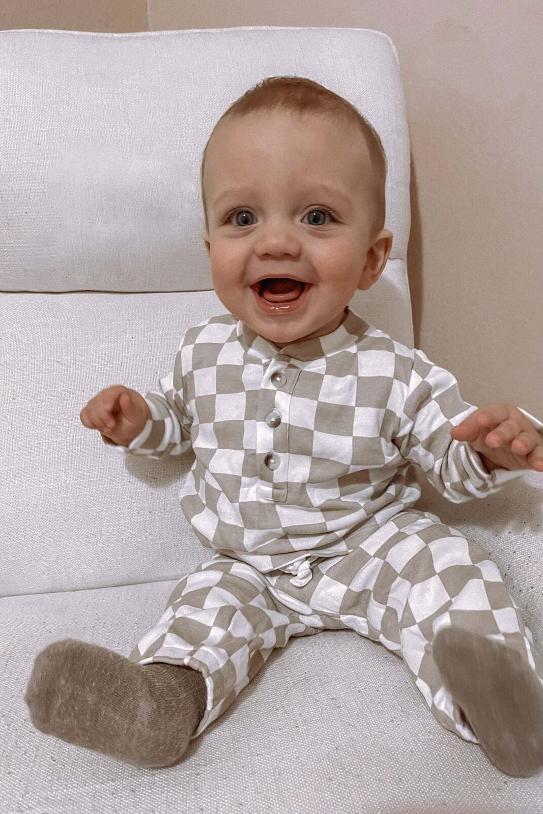 Smiling baby in a checkered outfit sitting on a white chair with excitement.