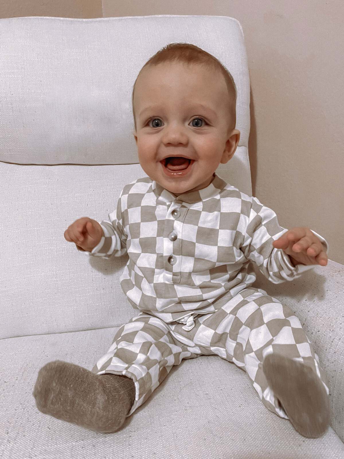 Smiling baby in a checkered outfit sitting on a white chair with excitement.