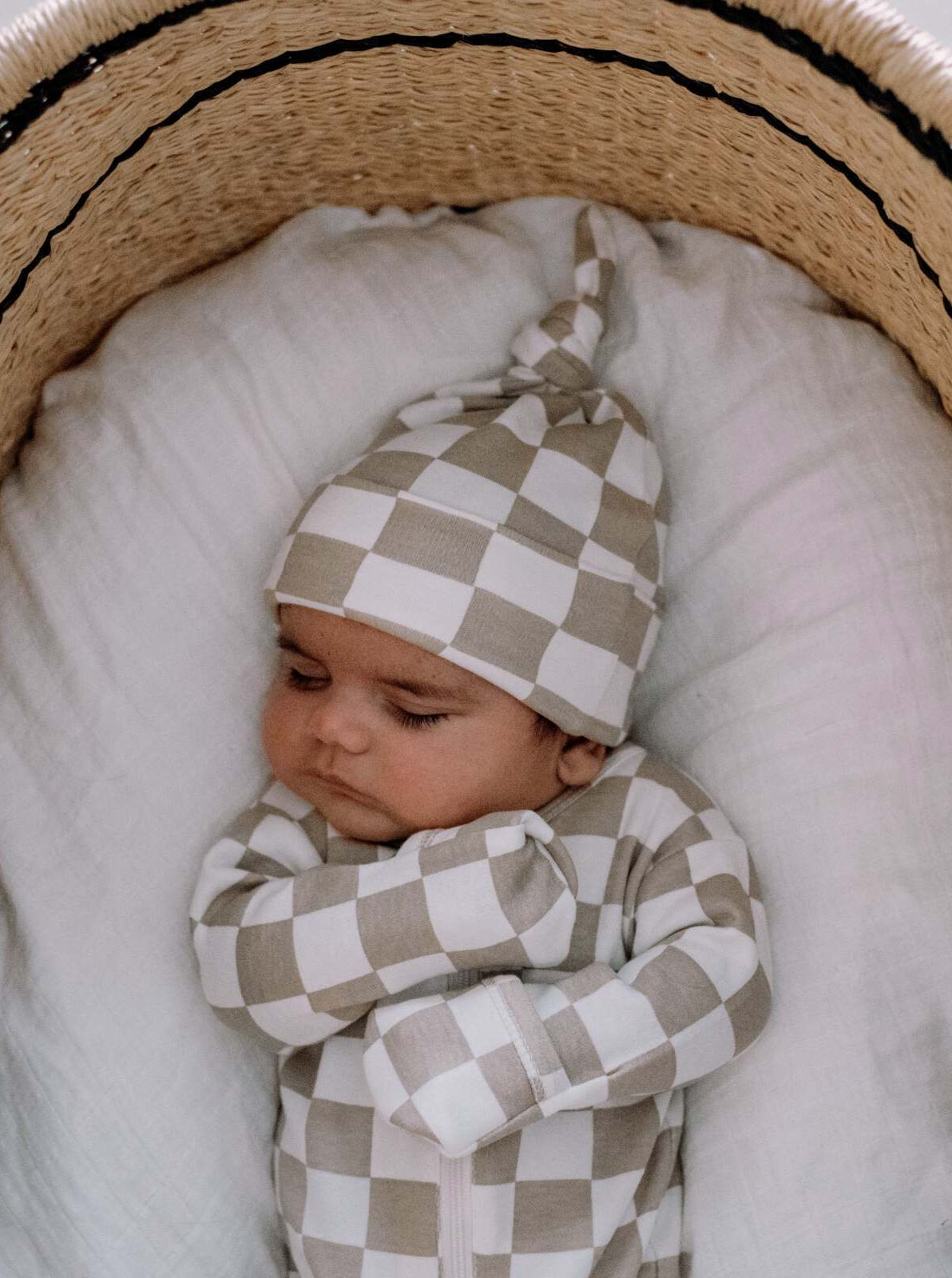 Sleeping baby in a checkered outfit, nestled in a cozy basket with a soft white blanket.