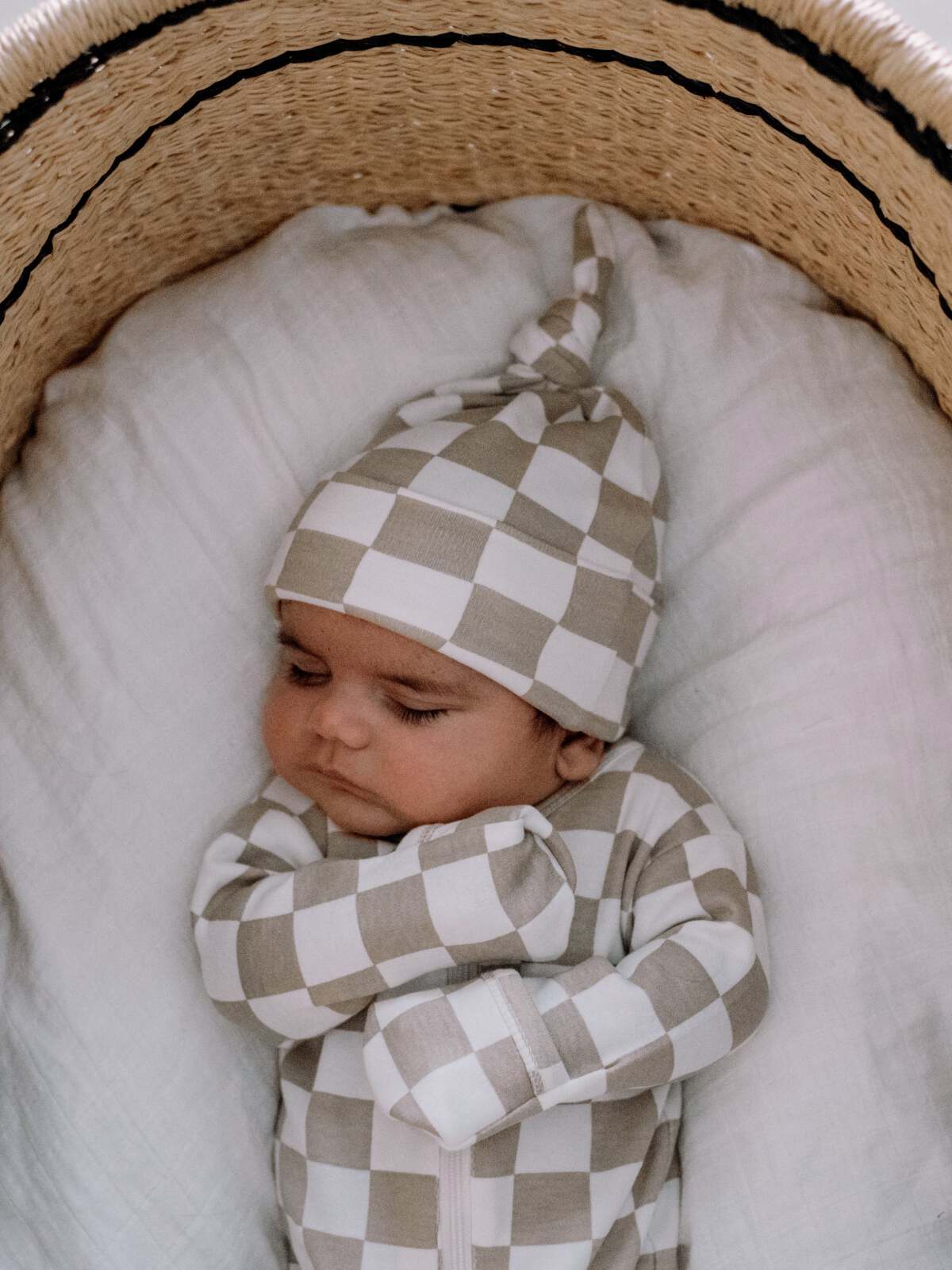 Sleeping baby in a checkered outfit, nestled in a cozy basket with a soft white blanket.