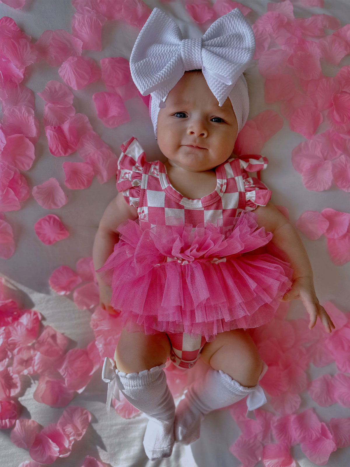 Baby in a pink tutu and oversized bow, lying on white fabric surrounded by pink flower petals.