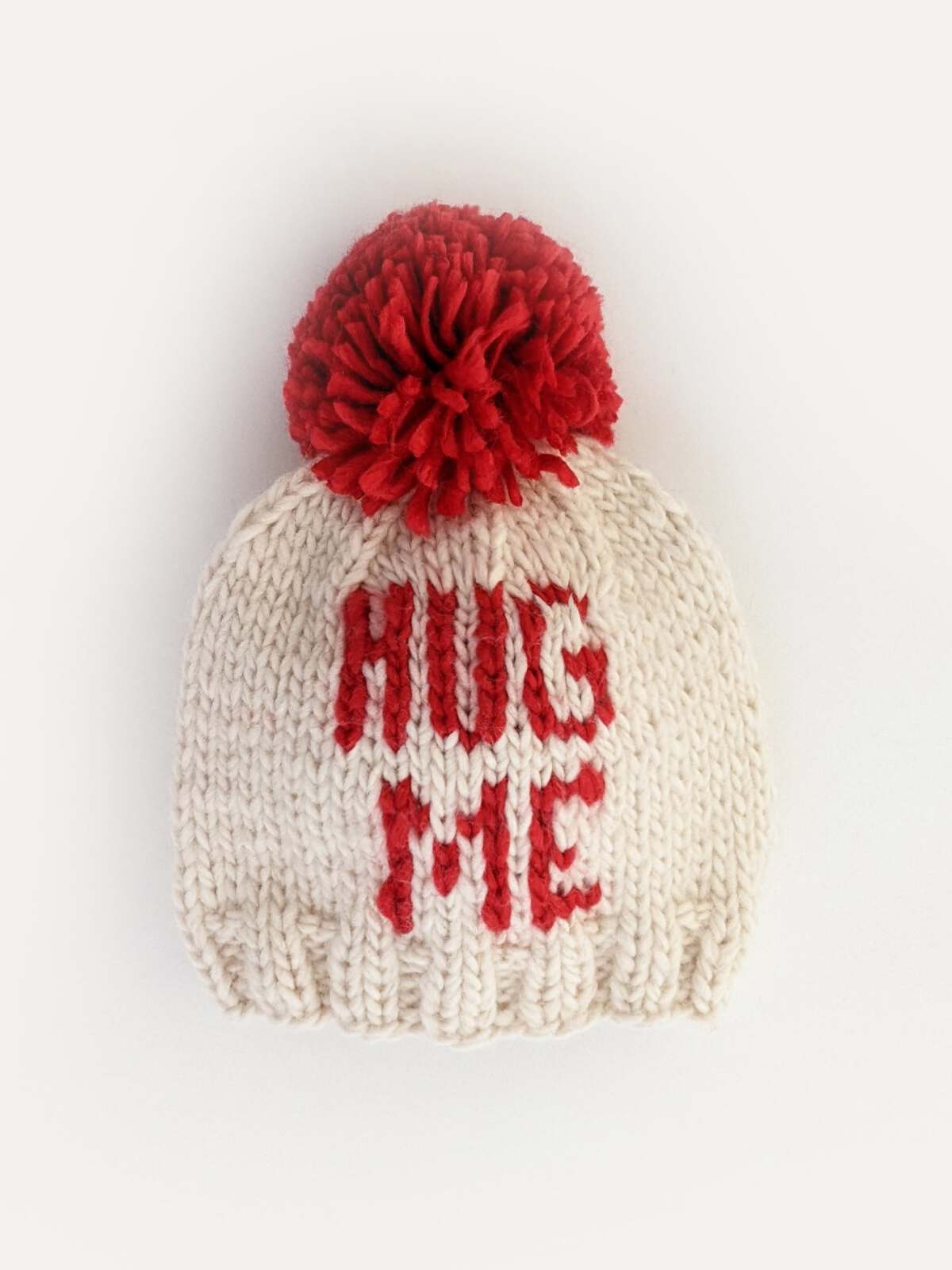 White knit beanie with a red pompom and "HUG ME" text on the front.