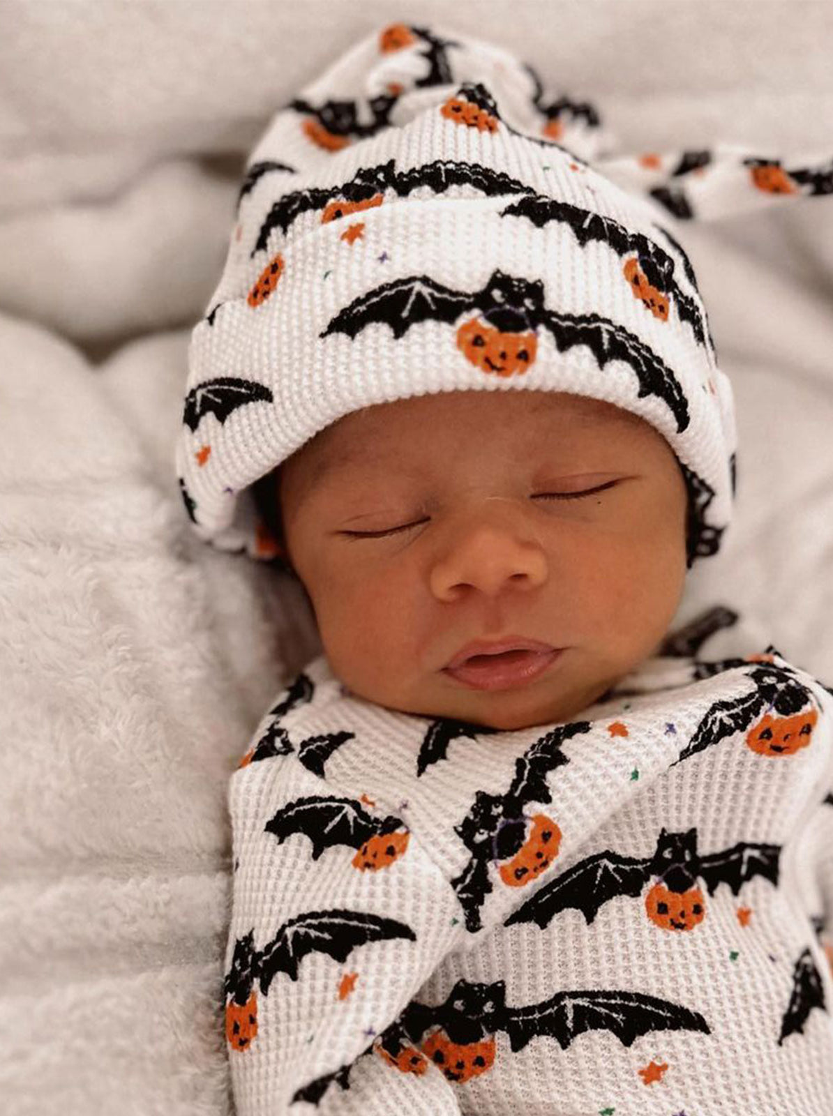 Sleeping baby wrapped in a bat and pumpkin-patterned blanket, wearing a matching hat.