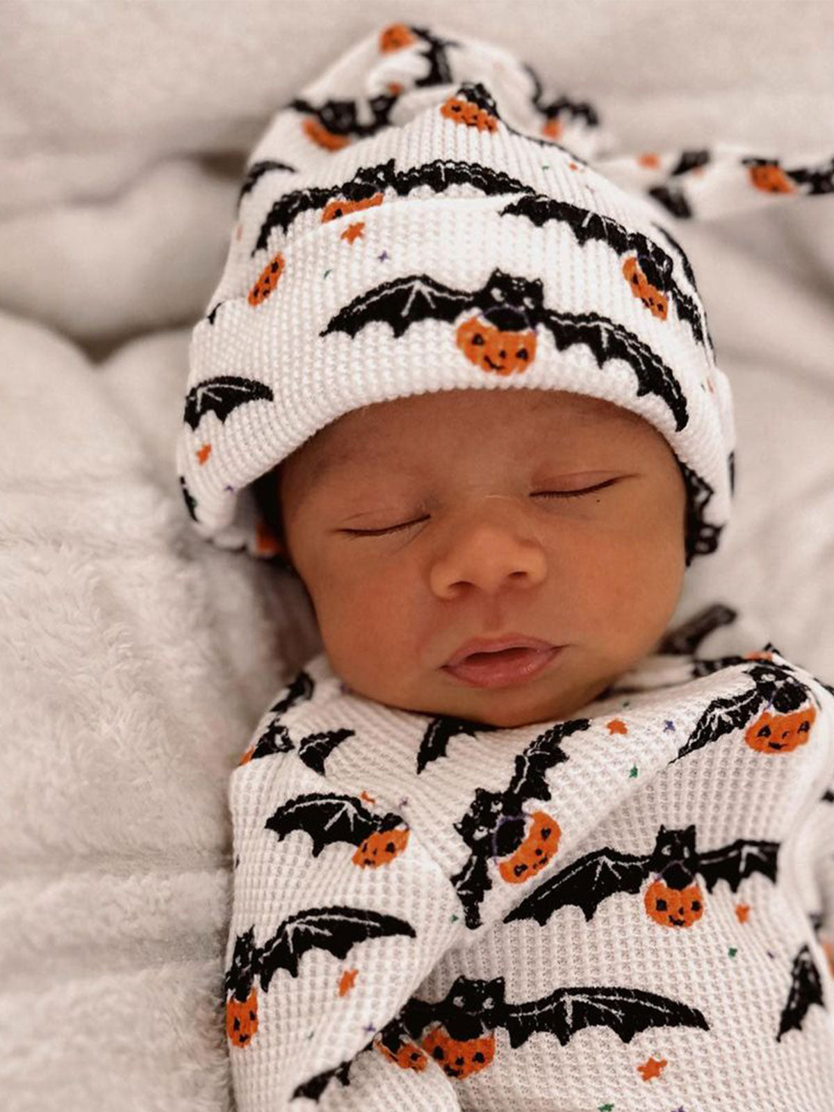 Sleeping baby wrapped in a bat and pumpkin-patterned blanket, wearing a matching hat.