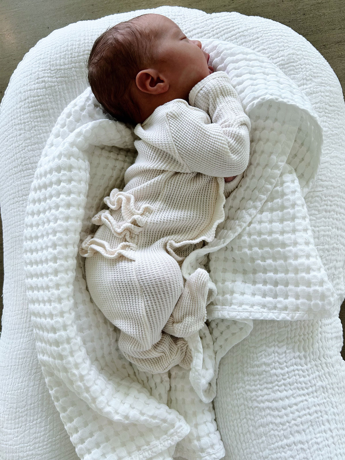 Newborn baby peacefully sleeping in a cozy white blanket and outfit, on a soft textured surface.