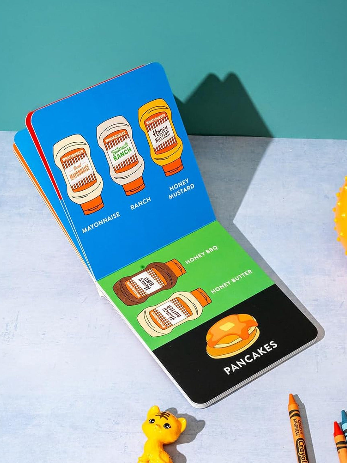 Colorful children's book page showing condiments and pancakes with a toy cat and crayons nearby.
