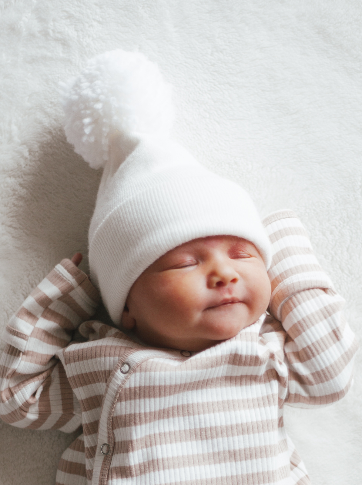 Cute baby sleeping on a soft blanket, wearing a white hat with a pom-pom and a striped outfit.
