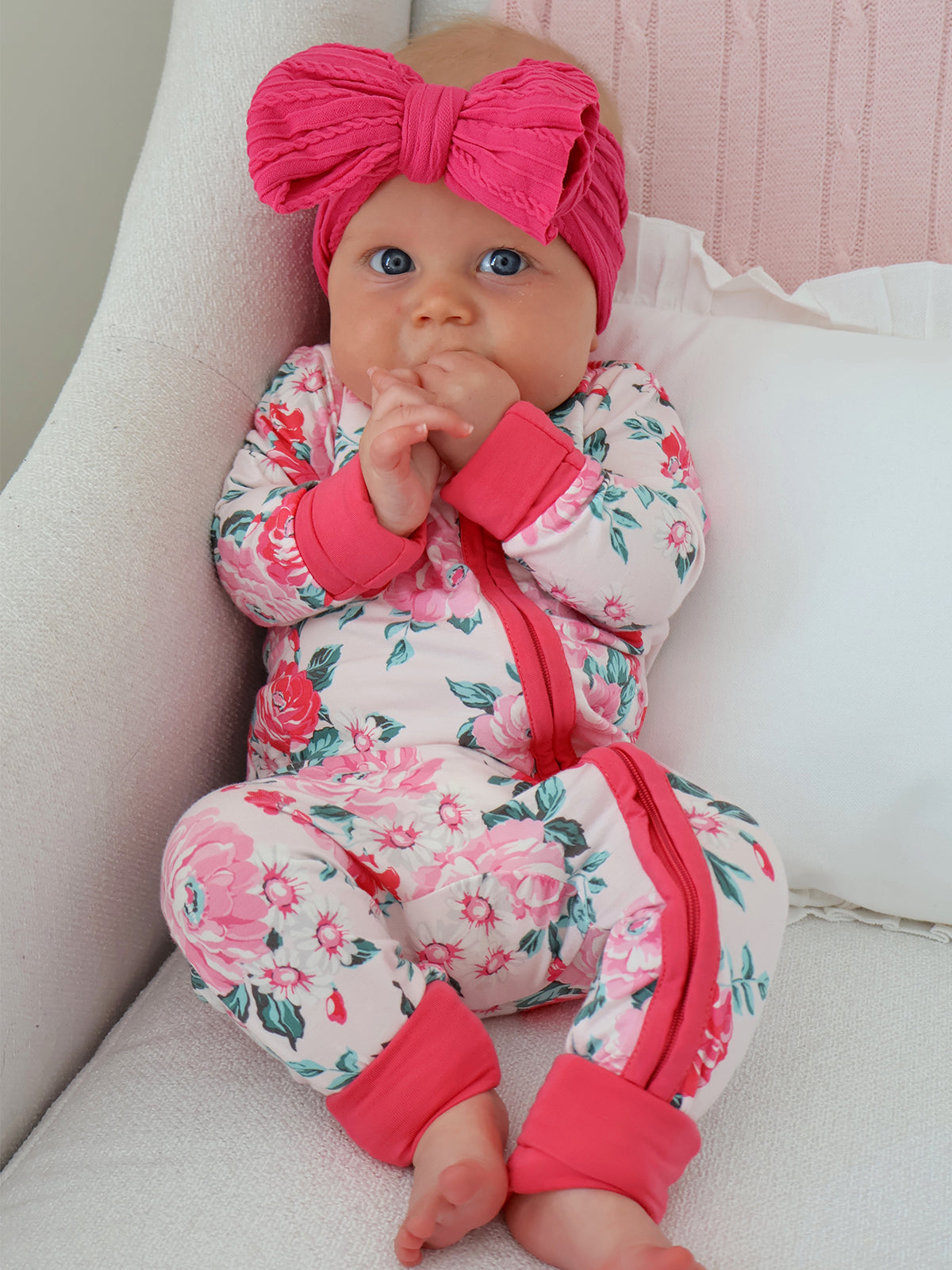 Infant in floral onesie with pink accents and large headband, sitting on a chair, looking curiously at the camera.