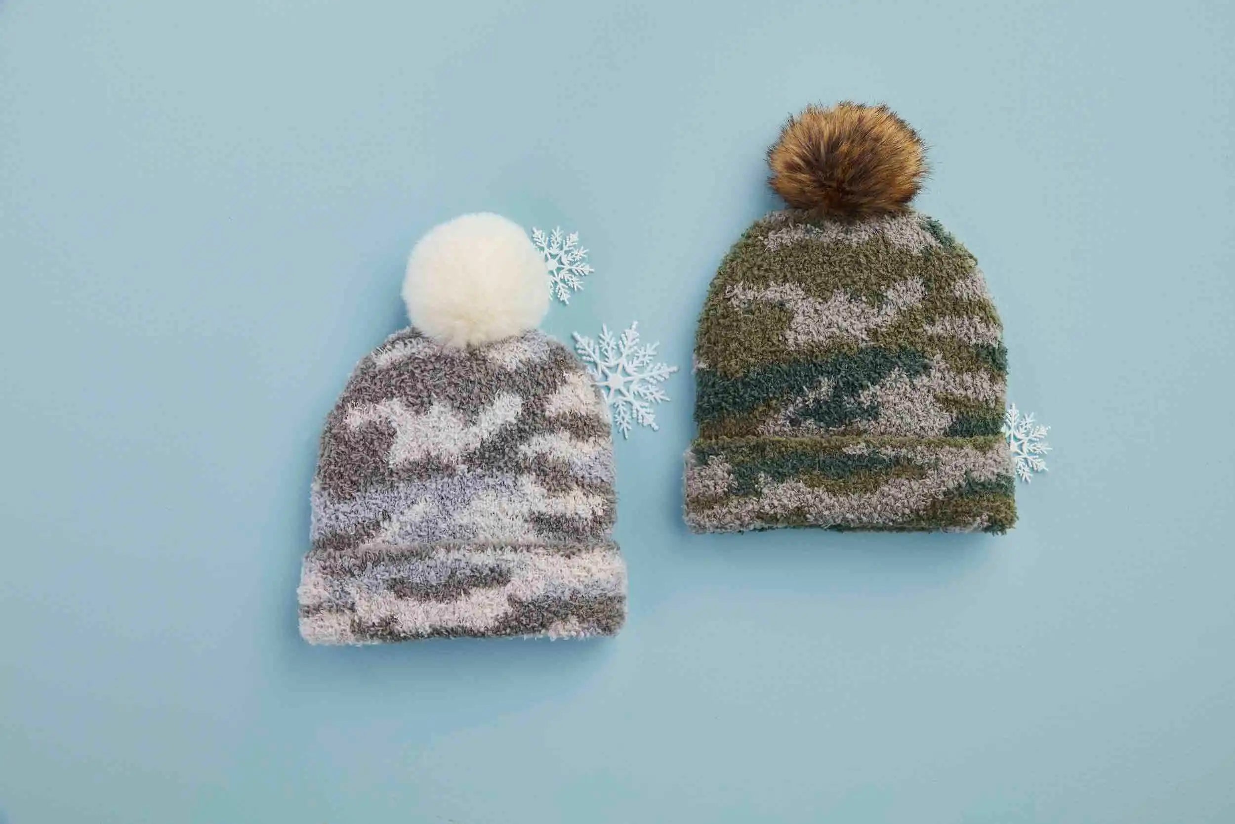 Two cozy winter beanies on a light blue background, one with a white pom-pom and the other with a brown pom-pom.