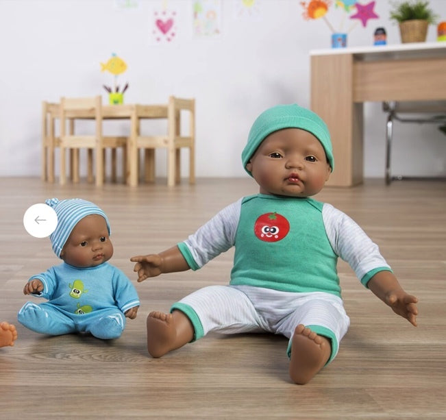 Two dolls sit on a floor, one wearing a blue outfit and the other in green. A colorful play area is visible in the background.