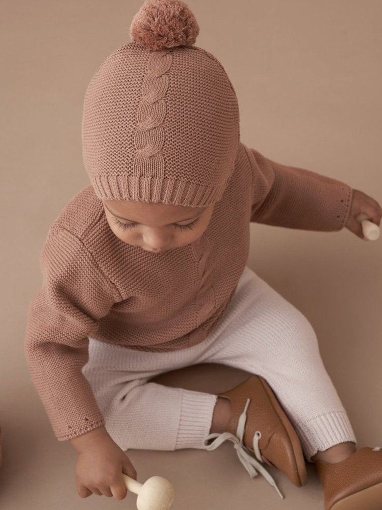 Baby wearing a knitted hat and sweater, sitting with a wooden toy and a basket on a soft, neutral background.