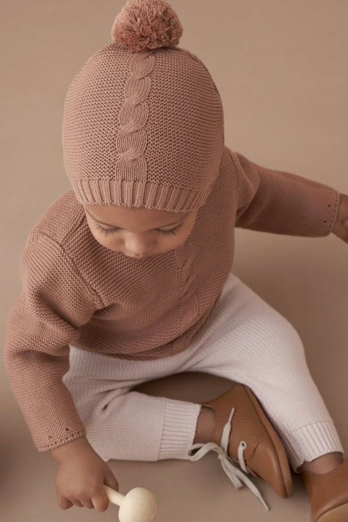 Baby wearing a knitted hat and sweater, sitting with a wooden toy and a basket on a soft, neutral background.