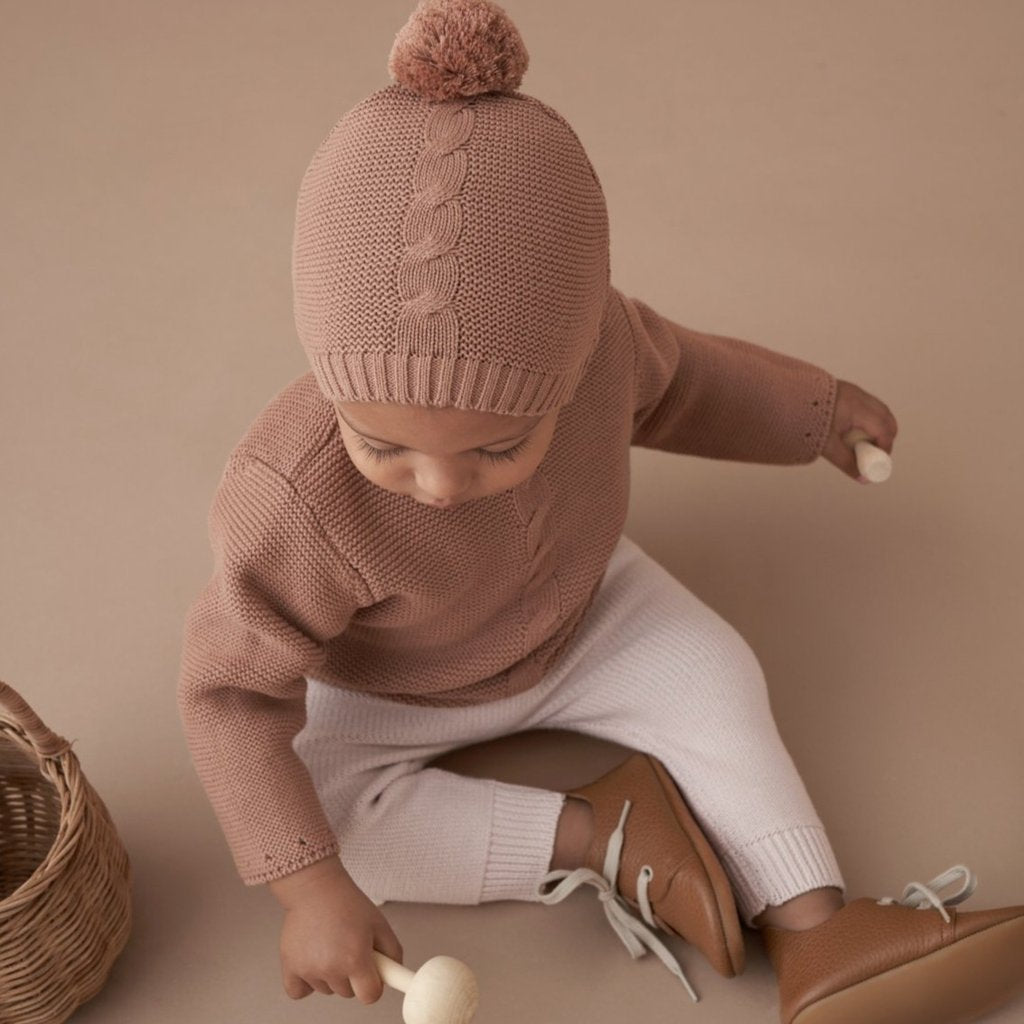 Baby wearing a knitted hat and sweater, sitting with a wooden toy and a basket on a soft, neutral background.