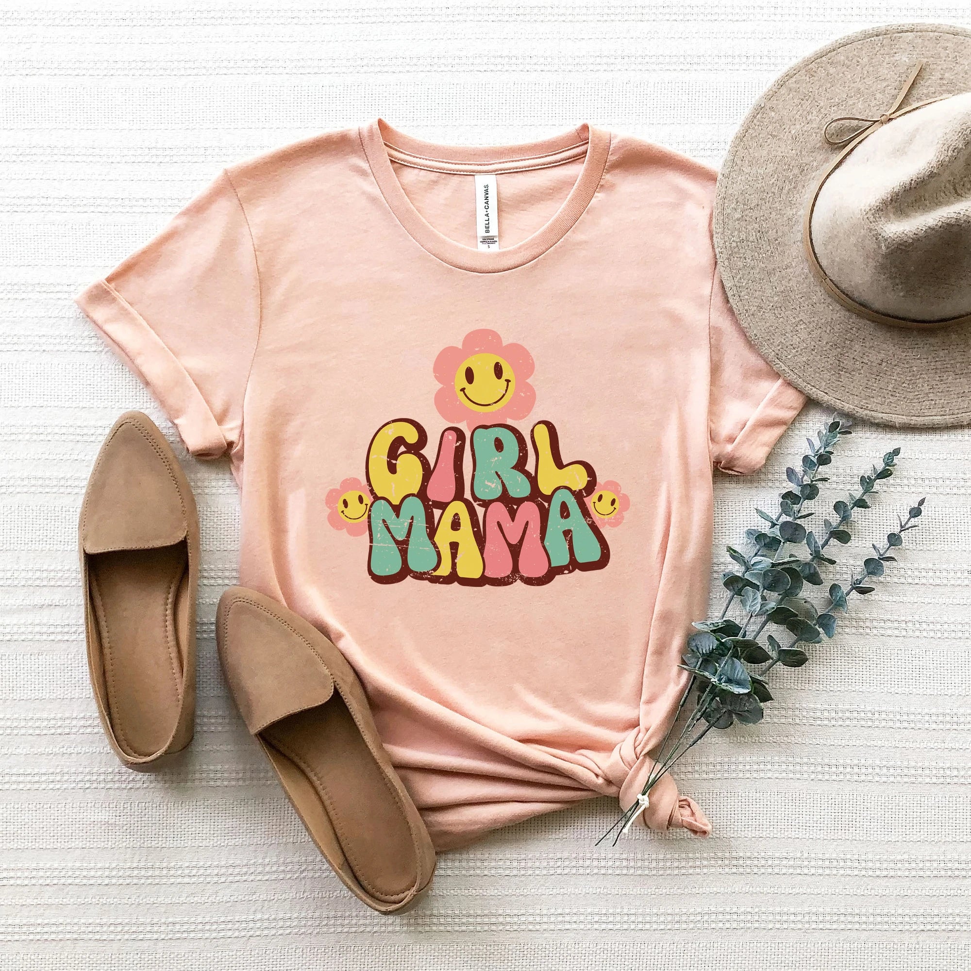 Peach t-shirt with "GIRL MAMA" design, accompanied by flats, a hat, and greenery, on a textured surface.