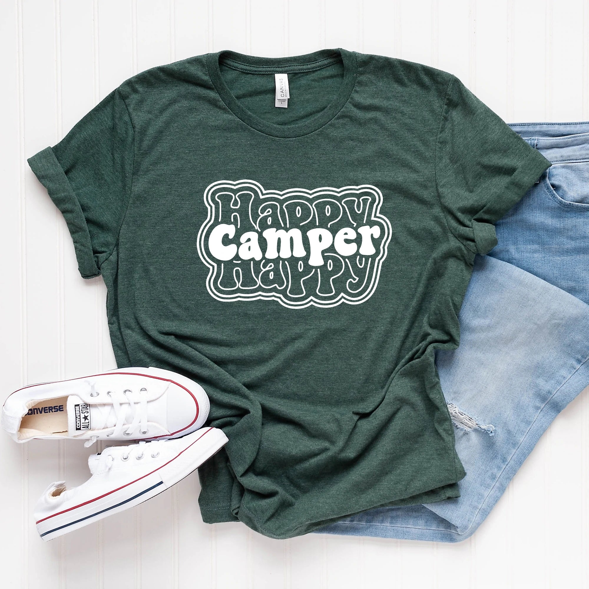 Green t-shirt with "Happy Camper Happy" graphic, paired with white sneakers and light jeans on a white background.