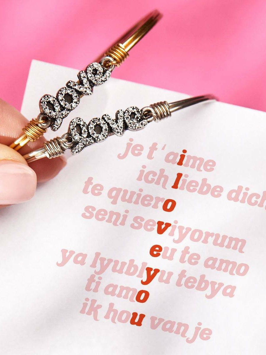 Hand holding two bracelets spelling "love," with multilingual phrases on a white sheet in the background.