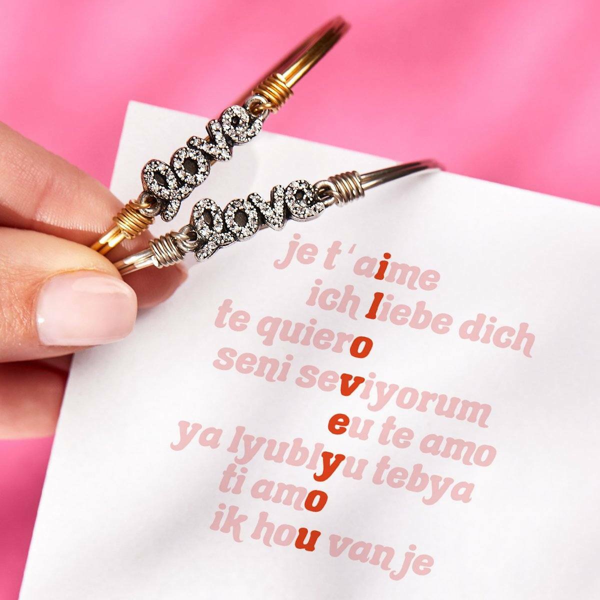 Hand holding two bracelets spelling "love," with multilingual phrases on a white sheet in the background.