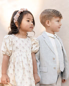Two children, a girl in a floral dress and a boy in a suit, standing side by side against a neutral background.
