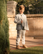 Smiling boy in a gray suit stands by a stone wall, showcasing stylish attire in a sunny outdoor setting.