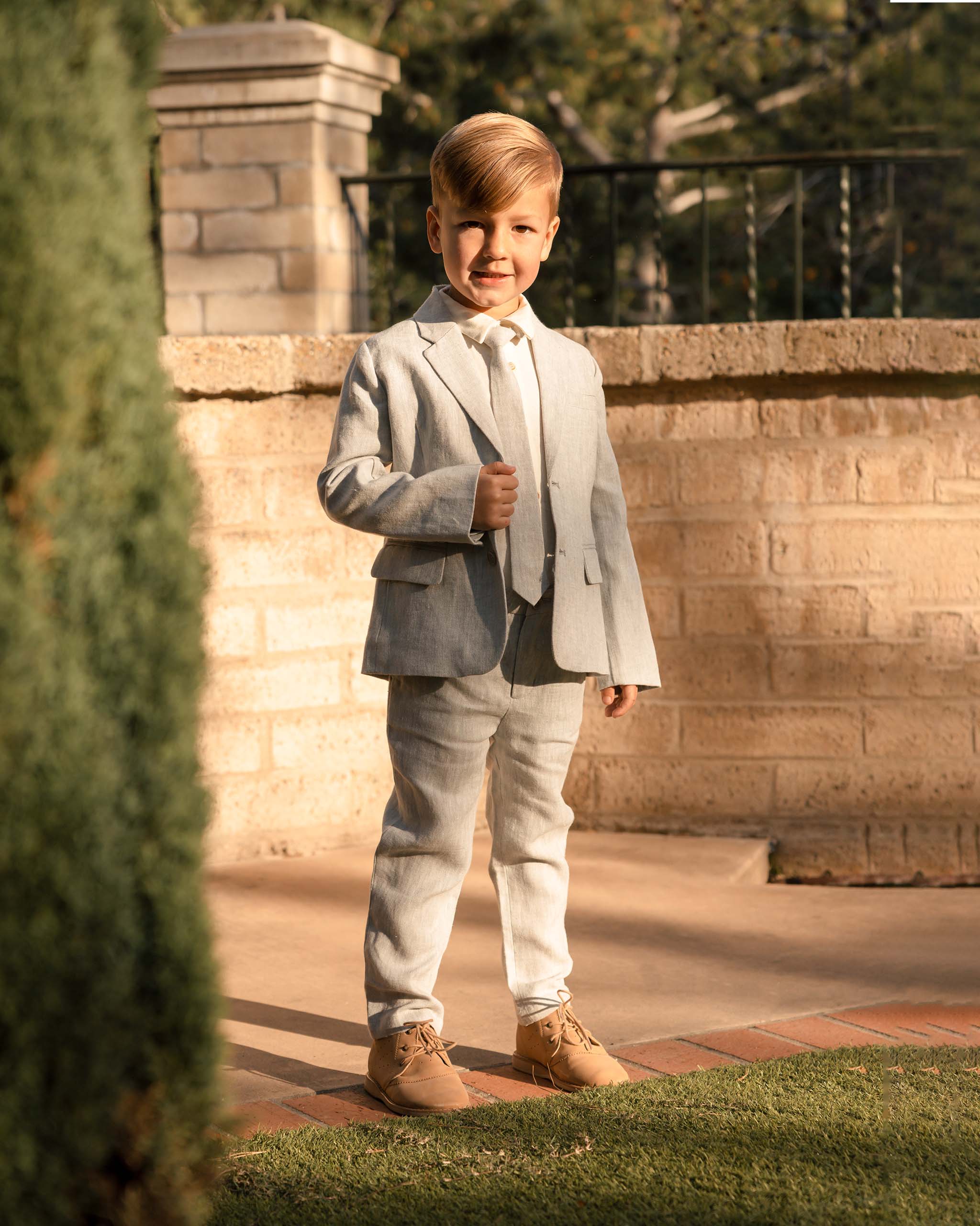 Smiling boy in a gray suit stands by a stone wall, showcasing stylish attire in a sunny outdoor setting.