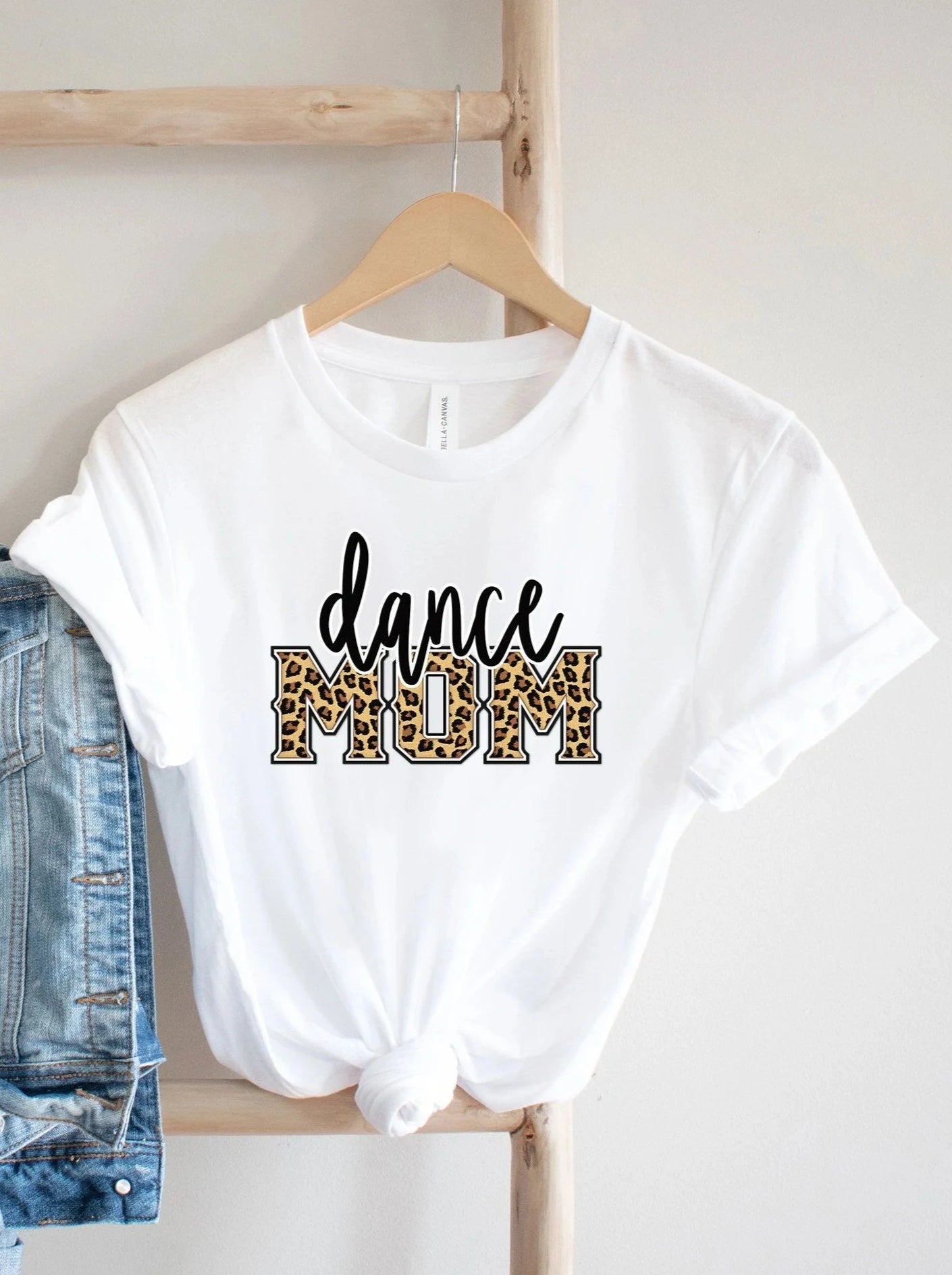 White t-shirt featuring "dance MOM" text with a leopard print design, displayed on a wooden hanger.