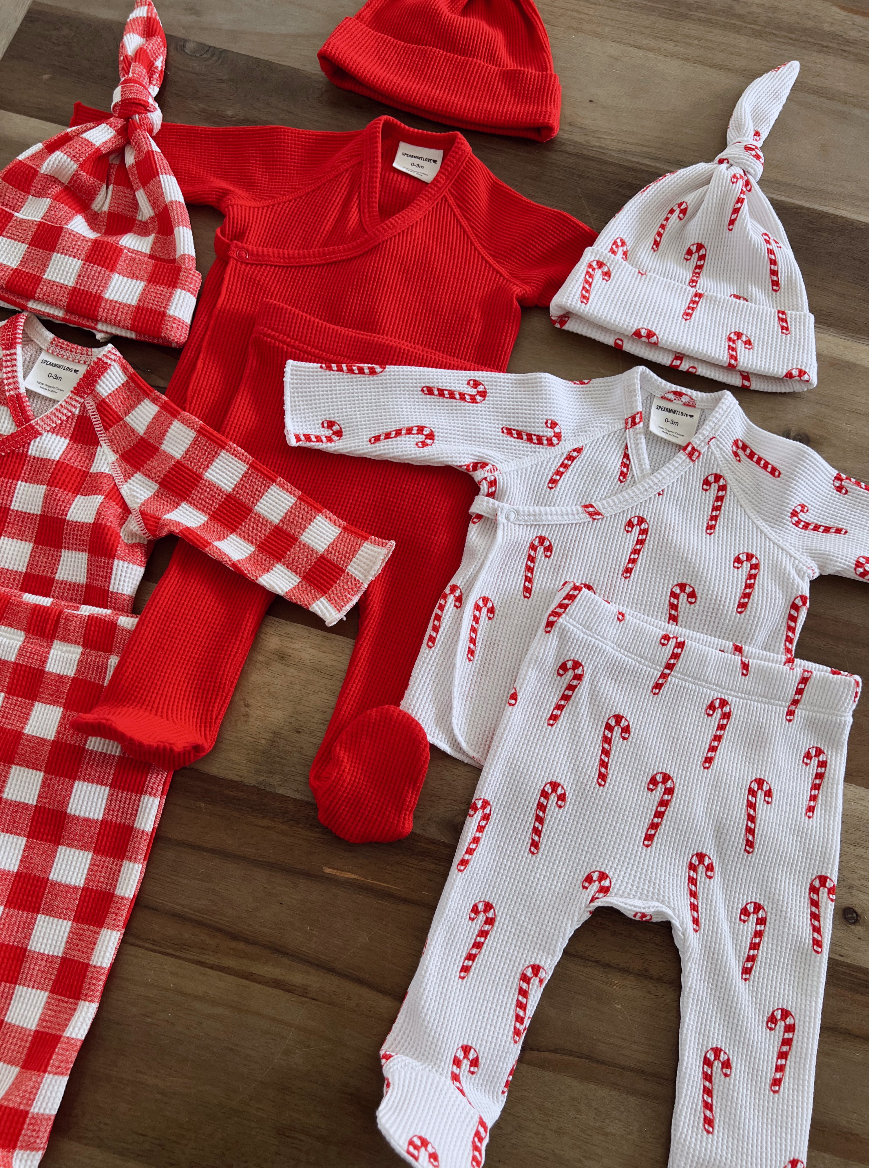 Colorful holiday baby clothes including red and candy cane patterned outfits and hats on a wooden surface.