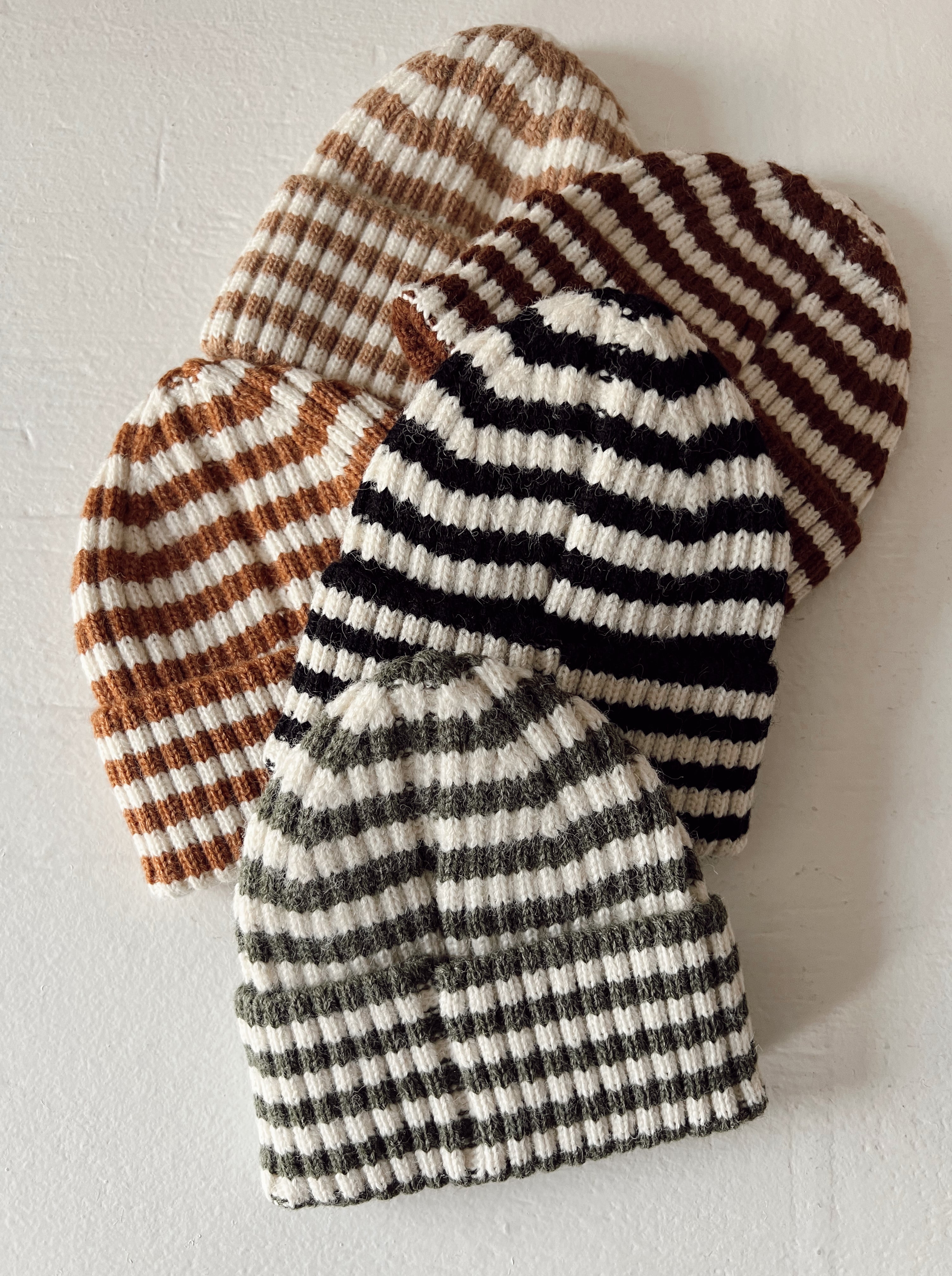 Four striped knit beanies in brown, black, and olive green stacked on a light surface.