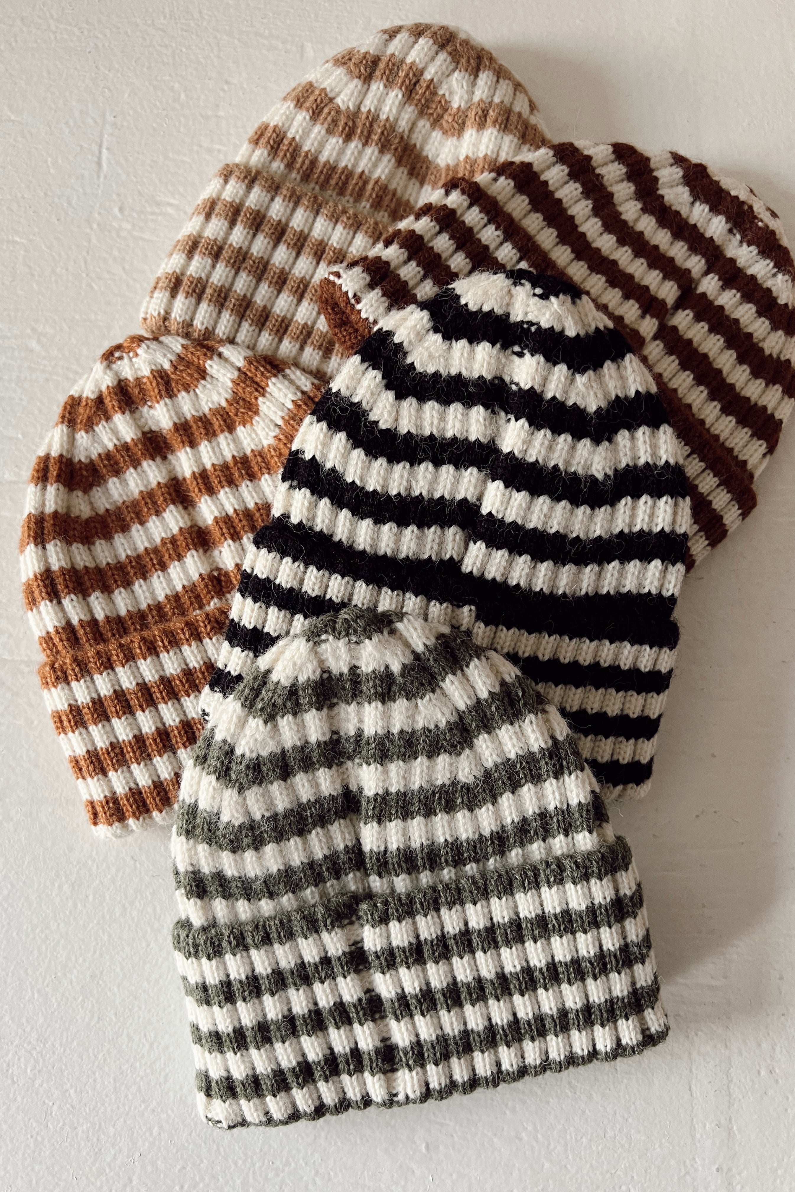 Four striped knit beanies in brown, black, and green, arranged on a light surface.