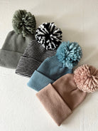 Four knitted beanies with playful pom-poms in gray, striped black and white, blue, and light pink.