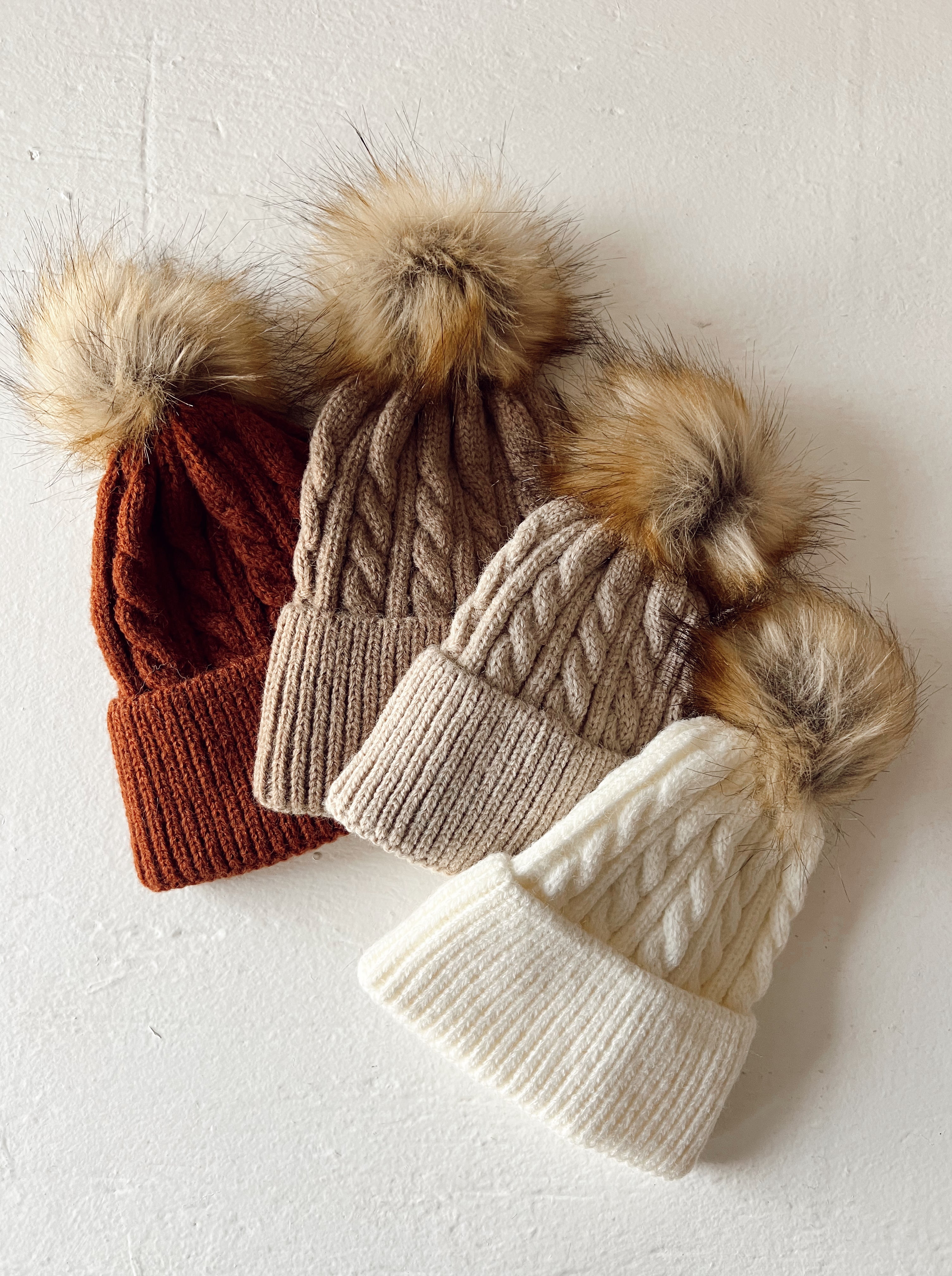 Three cozy knit beanies in rust, beige, and white with fluffy pom-poms on top against a textured white background.