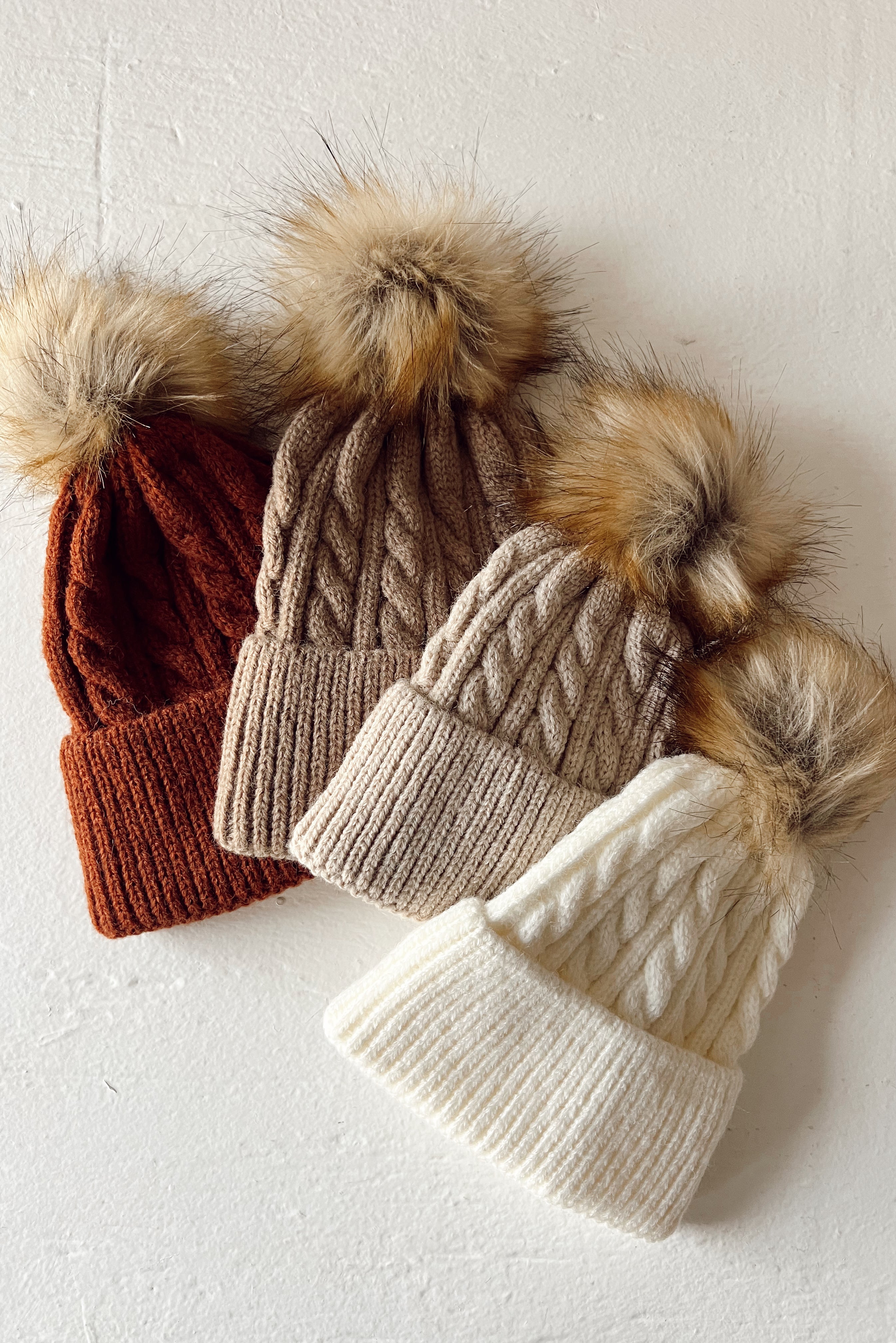 Three knitted beanies in rust, beige, and white, each with a fluffy pom-pom on top, against a white background.