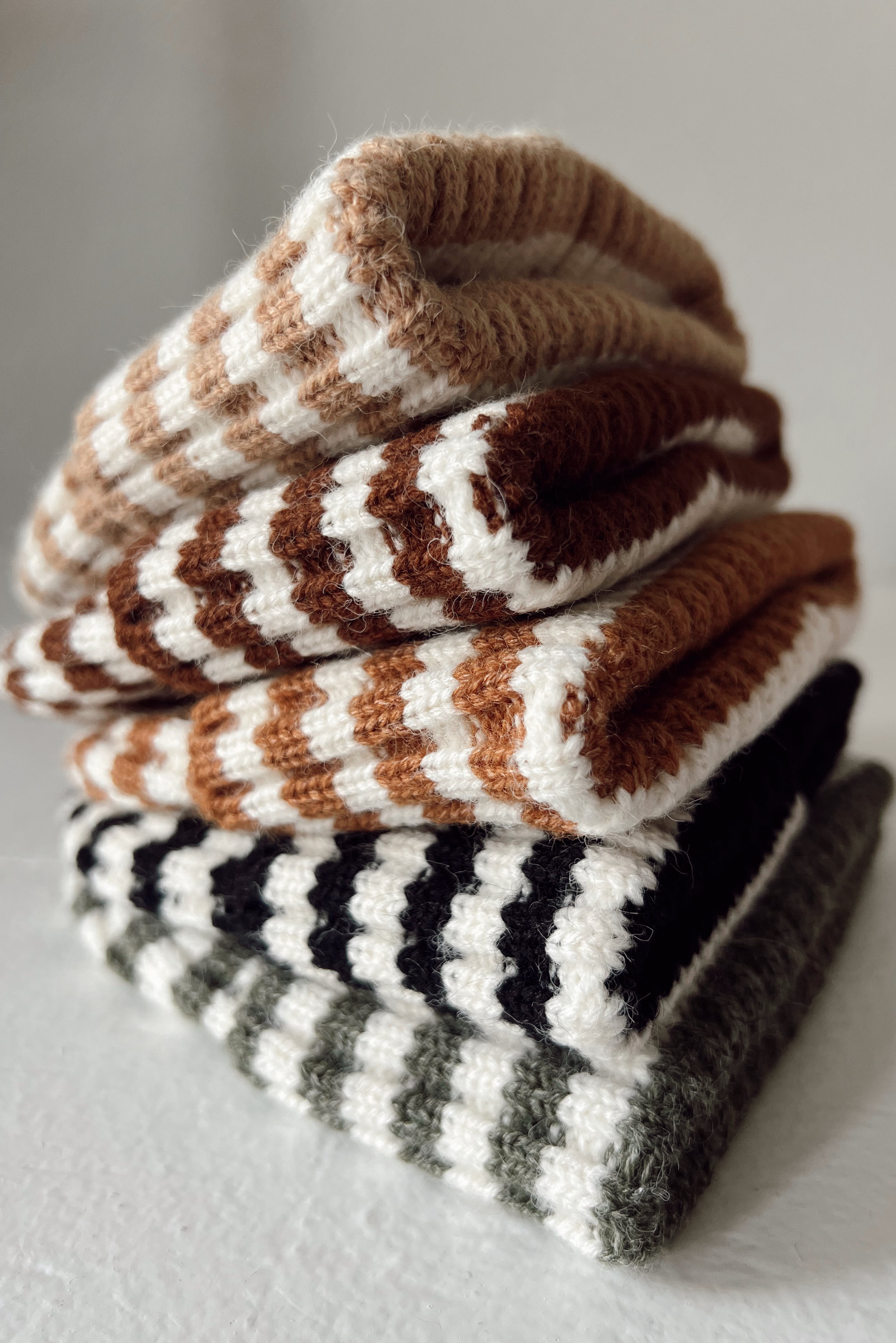 Stack of knitted, striped socks in earthy tones of brown, black, and green, resting on a textured surface.