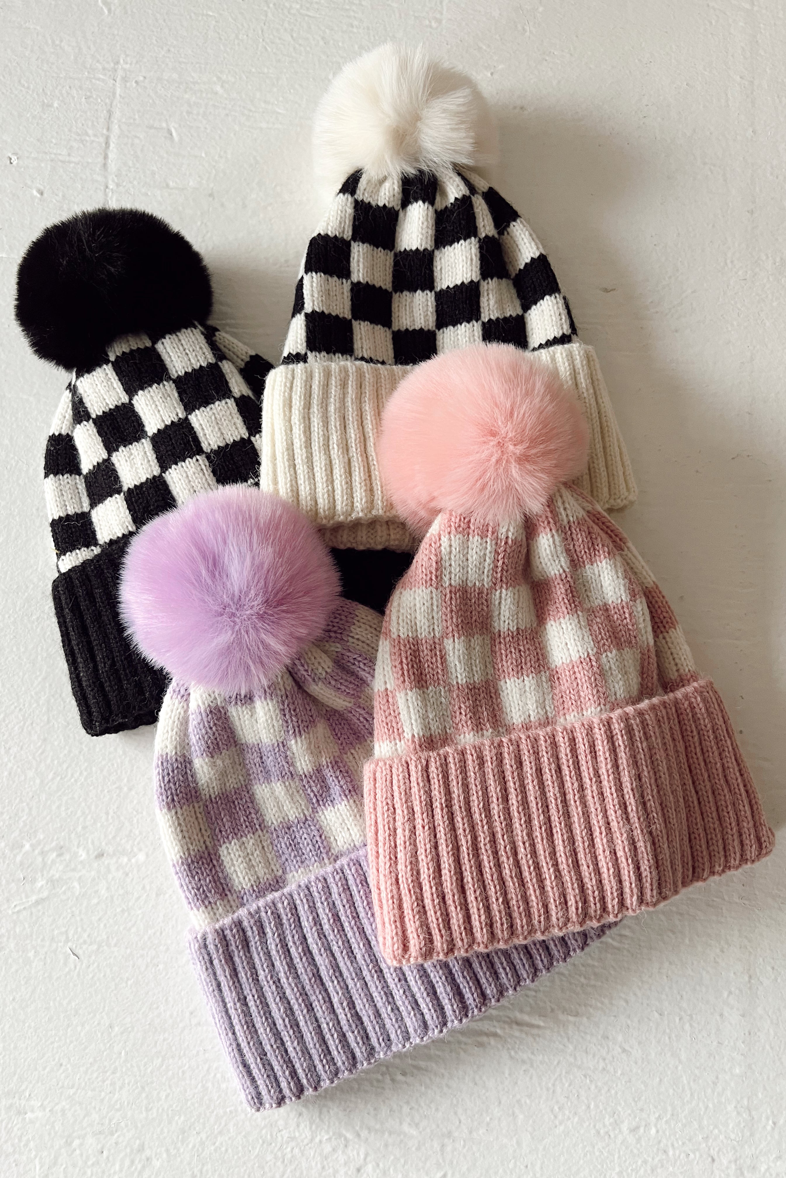 Four stylish beanies featuring pom-poms and checker patterns in various colors: black, pink, and lavender.