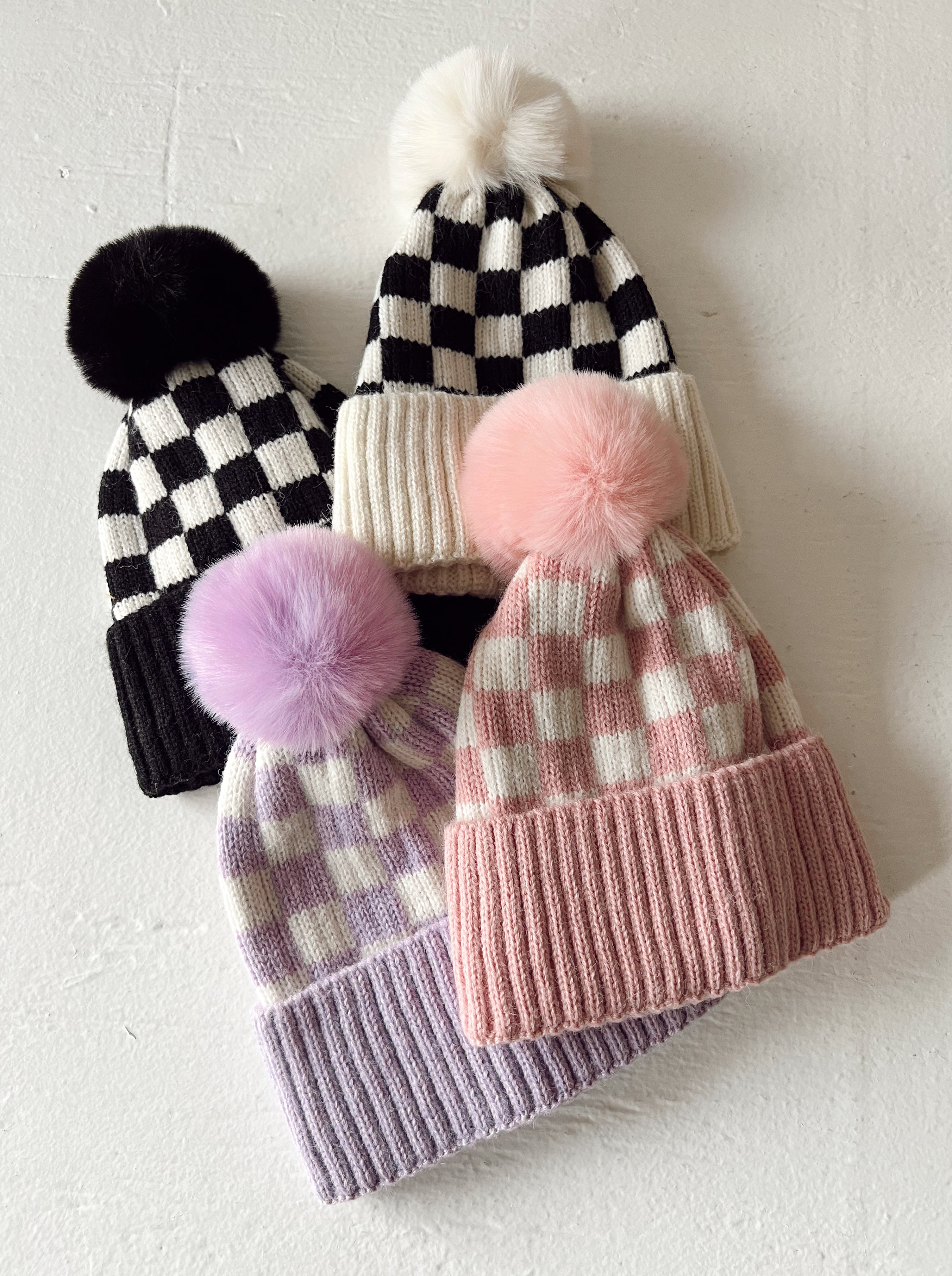 Four stylish beanies featuring pom-poms and checker patterns in various colors: black, pink, and lavender.